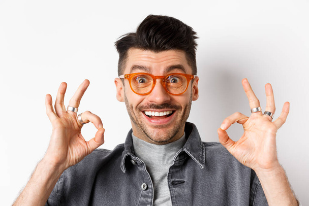 Primer plano retrato de los clientes masculinos satisfechos alabando las nuevas gafas, mostrando signo bien y sonriendo feliz, de pie sobre fondo blanco - Foto, imagen