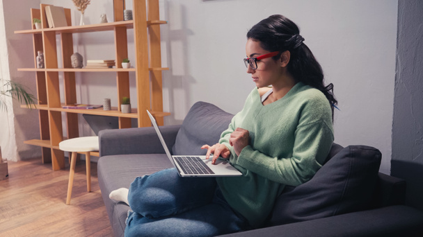 Боковой вид женщины в очках, работающей на ноутбуке дома  - Фото, изображение