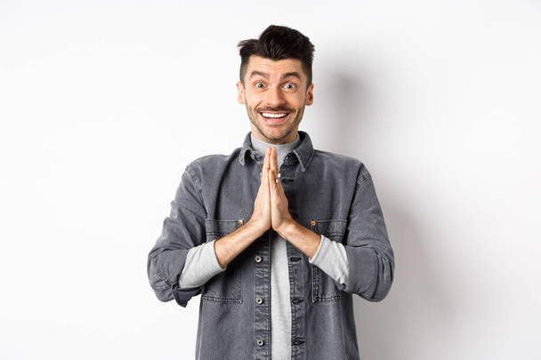 Please help me. Smiling cute man holding hands in namaste gesture, asking for favour, saying thank you, looking hopeful and excited at camera, standing on white background - Photo, Image