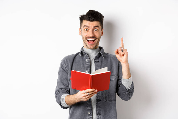 Excited guy pitching an idea while doing homework, raising finger and holding planner, standing against white background - Фото, зображення