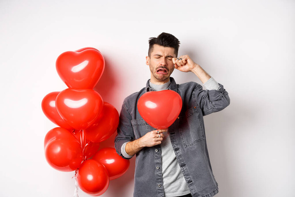 Valentines day and love concept. Sad crying man holding red heart balloon and whiping tears, standing single and miserable, being heartbroken, white background - Photo, Image