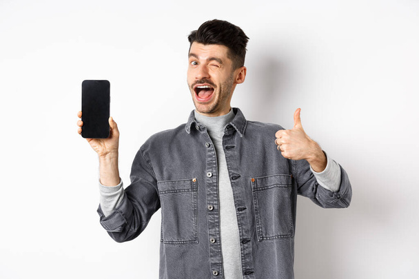Jóképű srác kacsint, és mutatja üres mobiltelefon képernyő hüvelykujjával felfelé, ajánló alkalmazás vagy online üzlet, álló ellen fehér háttér - Fotó, kép
