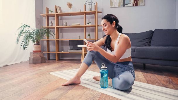 Positive woman in sportswear using smartphone near sports bottle at home  - Photo, Image
