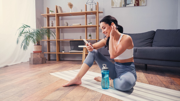 Seitenansicht der Sportlerin beim Videochat auf dem Smartphone, während sie zu Hause auf der Fitnessmatte neben der Sportflasche sitzt  - Foto, Bild
