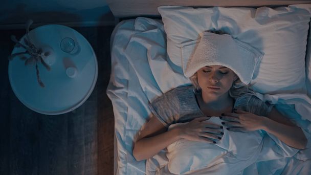 Vista superior de la mujer enferma acostada en la cama cerca del agua y pastillas en el dormitorio  - Foto, imagen