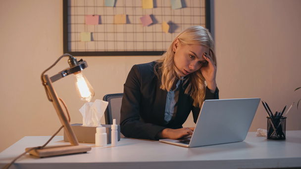 Femme d'affaires épuisée utilisant un ordinateur portable près de pilules, serviette et pulvérisation nasale au bureau - Photo, image