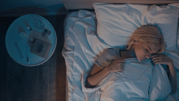 Vista superior de la mujer durmiendo cerca de píldoras, agua y aerosol nasal en la mesita de noche  - Foto, Imagen