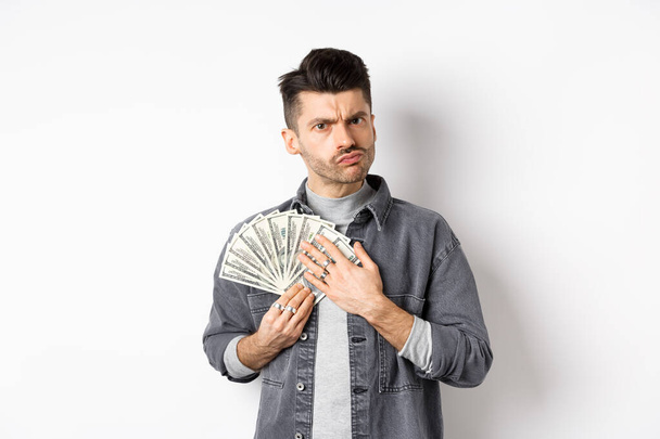 Άπληστος τύπος κατσουφιάζει και αγκαλιάζει χαρτονομίσματα, απρόθυμος να μοιραστεί χρήματα, στέκεται σε λευκό φόντο - Φωτογραφία, εικόνα