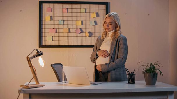 妊娠中のビジネスマン夜のオフィスでラップトップと水のガラスの近くに腹を触れる女性  - 写真・画像