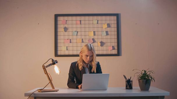 Businesswoman using laptop near lamp and stationery in office in evening  - Foto, afbeelding