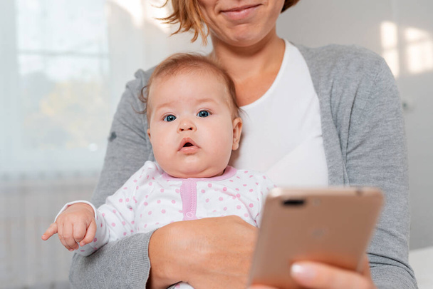 Portrait of a baby sitting in the arms of a smiling mother who uses a smartphone. Bottom view. Smartphone is blurred in the foreground. - Photo, Image