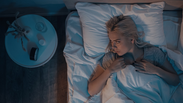 Draufsicht einer Frau mit Schlaflosigkeit beim Blick auf den Wecker in der Nähe von Tabletten im Schlafzimmer  - Foto, Bild