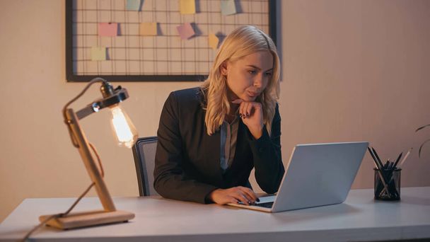 Businesswoman working on laptop near lamp in office in evening  - Zdjęcie, obraz