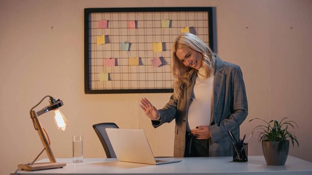 Uśmiechnięta ciężarna bizneswoman machająca ręką na laptopie w biurze  - Zdjęcie, obraz