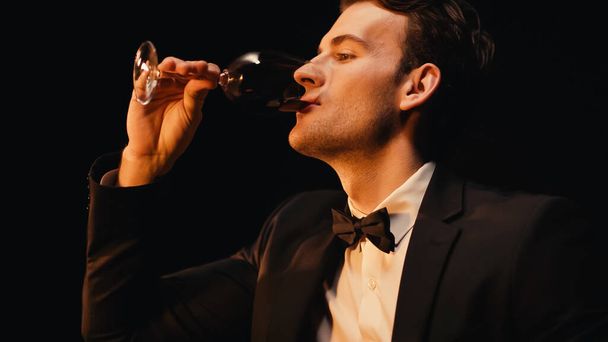Pewny siebie młody człowiek w garniturze pijący czerwone wino odizolowane na czarno  - Zdjęcie, obraz