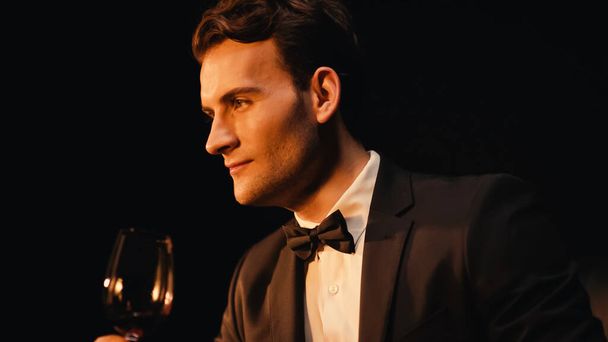 Уверенный молодой человек в костюме держит бокал красного вина изолирован на черном  - Фото, изображение