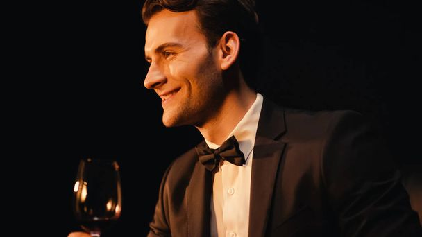 boldog fiatalember öltönyben kezében pohár vörösbor elszigetelt fekete  - Fotó, kép
