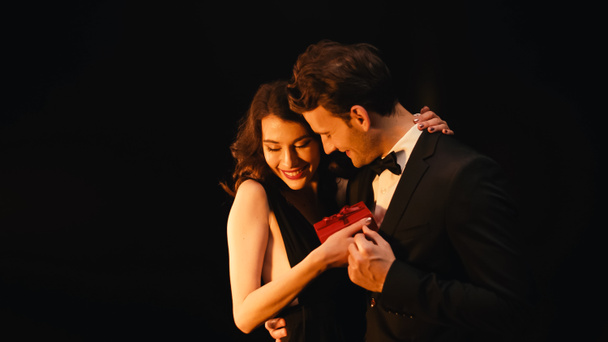 szczęśliwy młody kobieta otrzymywać prezent pudełko i przytulanie wesoły mężczyzna w garniturze odizolowany na czarny  - Zdjęcie, obraz