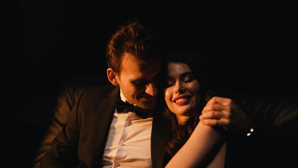 Hombre sonriente en traje abrazando y coqueteando con novia complacida aislada en negro  - Foto, imagen
