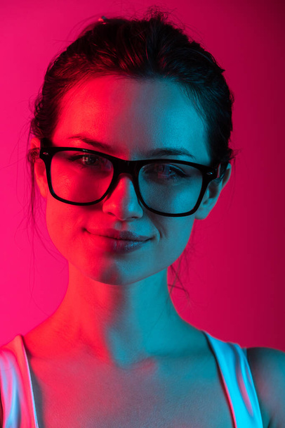 Close-up cropped portrait of young beautiful girl in glasses on gradient pink red background in neon lights - Zdjęcie, obraz