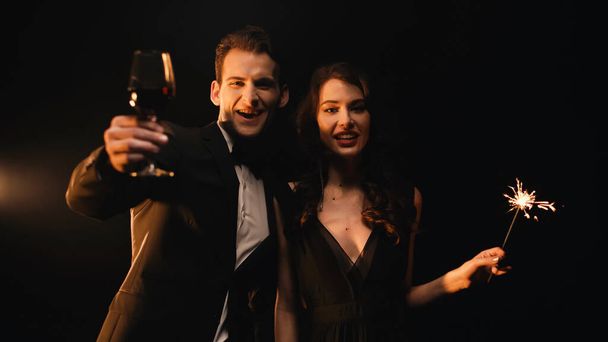 cheerful man holding glass of red wine and woman with sparkler looking at camera on black  - Photo, Image