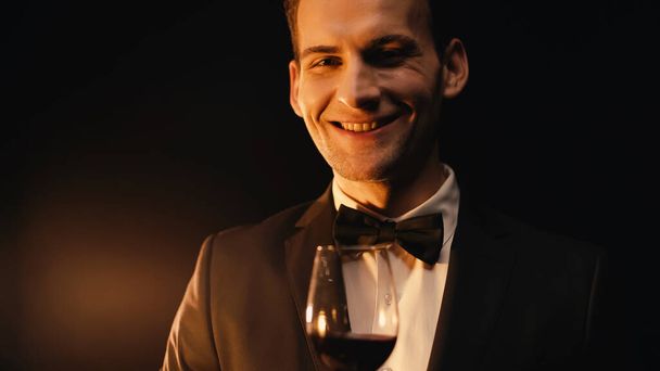 cheerful young man in suit with bow tie holding glass of red wine and looking at camera on black  - Foto, Imagem