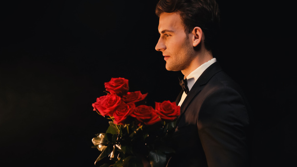 Vista lateral de un joven elegante sosteniendo rosas rojas aisladas en negro  - Foto, Imagen