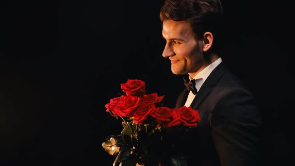 Vista lateral de un joven alegre sosteniendo rosas rojas aisladas en negro  - Foto, imagen