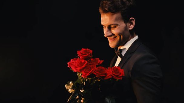 glücklicher junger Mann im Anzug mit roten Rosen auf schwarzem Hintergrund  - Foto, Bild