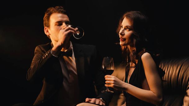 joven mujer sosteniendo vidrio mientras hombre beber vino tinto aislado en negro - Foto, imagen