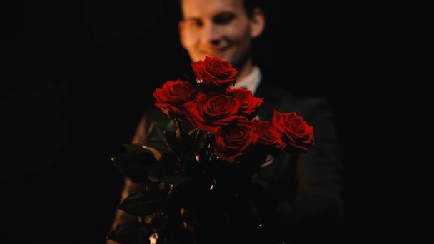 gelukkig en wazig man met rode rozen geïsoleerd op zwart  - Foto, afbeelding
