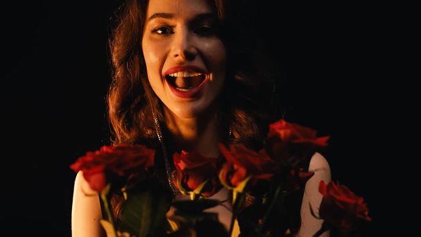 Portrait of happy woman with open mouth and red roses looking at camera isolated on black  - Fotografie, Obrázek