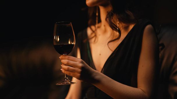 vista recortada de la elegante mujer joven sosteniendo vaso de vino y sentado en el sofá en negro  - Foto, imagen