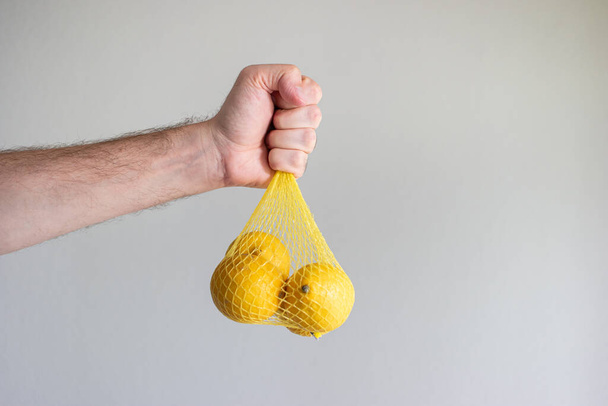 Свежие спелые желтые лимоны в мешке, подержанной за руку белой мужской рукой. Изолированная студия. - Фото, изображение