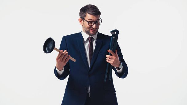 Cheerful businessman in suit holding pipe wrench and plunger isolated on white - Φωτογραφία, εικόνα