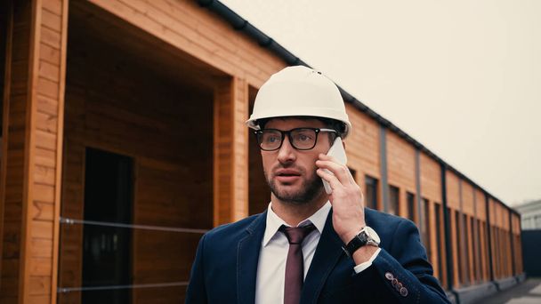 Engineer in white hardhat talking on mobile phone near building on urban street  - Foto, Bild