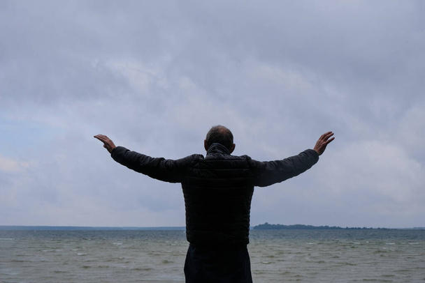Egy 60 éves férfi egy viharos tóparti háttérben olyan, mintha egy szimfónia karmestere lenne. Az eső után. Vicces fotó.. - Fotó, kép