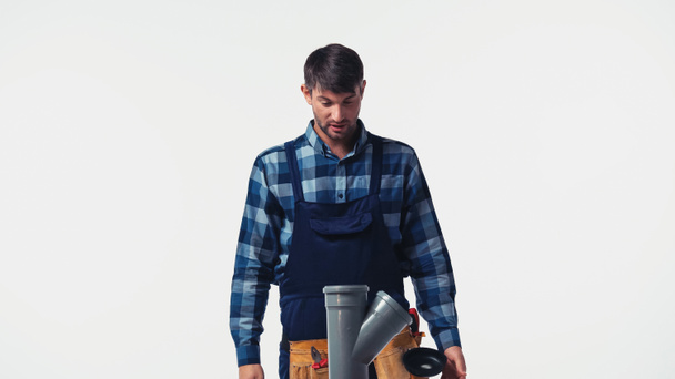 Trabajador en overoles y cinturón de herramientas mirando tubo de plástico aislado en blanco - Foto, imagen