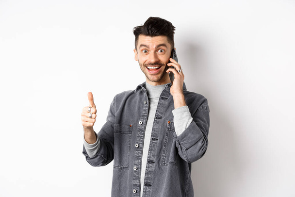 Heyecanlı genç adam telefonda konuşuyor ve baş parmağını kaldırıyor, gülümsüyor, beyaz arka plana karşı duruyor. - Fotoğraf, Görsel