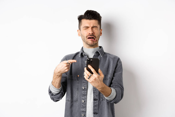 Sad crying guy pointing at smartphone and sobbing, complaining or feeling miserable, standing on white background - Фото, зображення