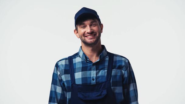 Cheerful handyman in overalls and cap looking at camera isolated on white - 写真・画像
