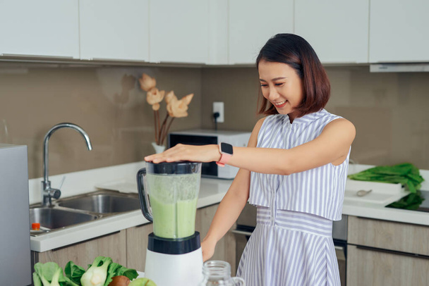 Giovane donna asiatica utilizzando frullatore per fare frullato verde in cucina
. - Foto, immagini