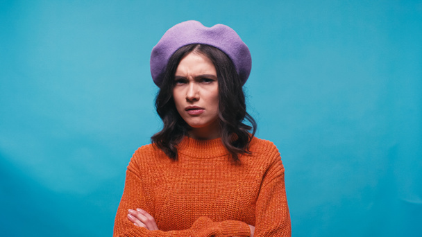 distrustful woman in purple beret and orange jumper looking at camera isolated on blue - Foto, imagen