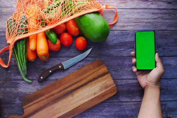 Blick von oben auf die Hand eines Mannes, der ein Smartphone mit frischem Gemüse auf dem Tisch hält  - Foto, Bild