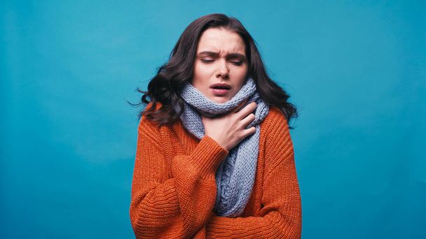 ill woman in warm scarf touching neck while suffering from pain in throat isolated on blue - Photo, Image