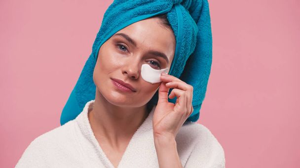 woman with blue towel on head removing eye patch isolated on pink - Фото, изображение