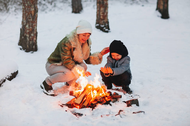 Wesoła matka i syn cieszą się gorącym napojem podczas odpoczynku przy ognisku wieczorem w zimowym lesie. Tydzień rodzinny - Zdjęcie, obraz