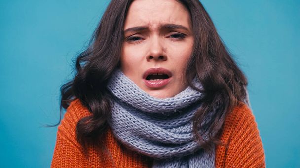 ill woman in knitted scarf sneezing while looking at camera isolated on blue - Foto, Imagen