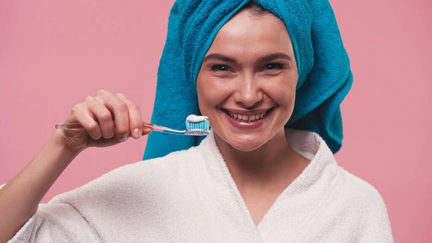 cheerful woman holding toothbrush with toothpaste while smiling at camera isolated on pink - Photo, Image
