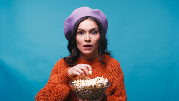impressed woman holding bowl of popcorn while watching interesting film isolated on blue - Photo, Image